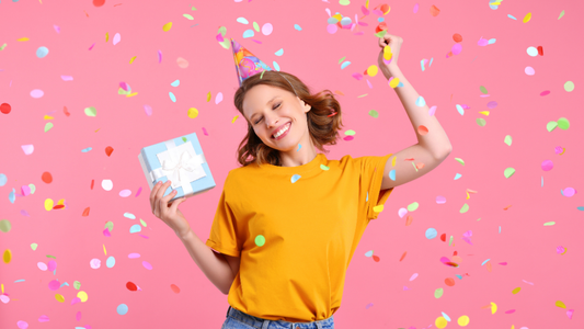 A lady holding a gift smiling and wearing a party hat with confetti falling around her
